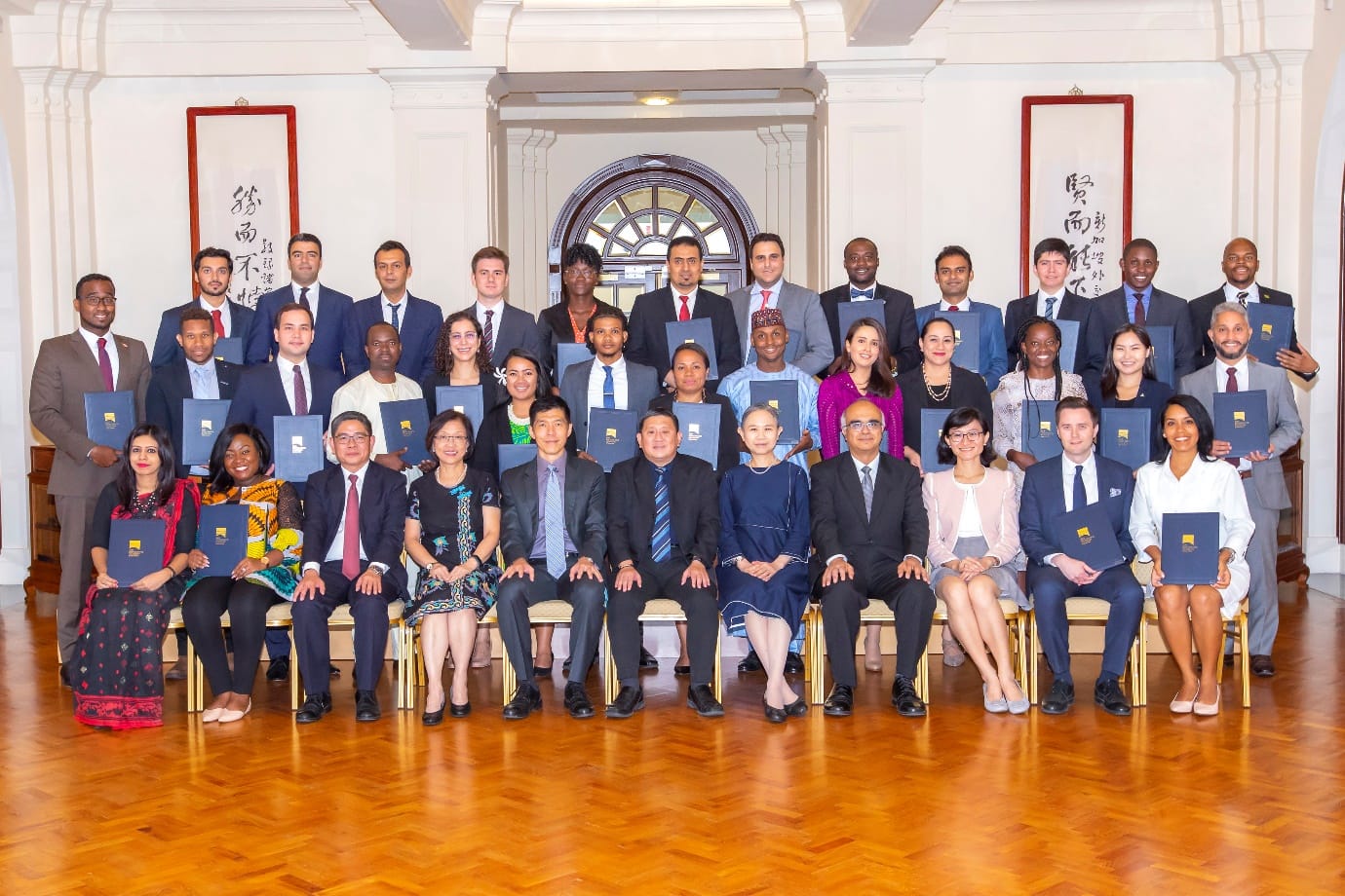 Group photo of diverse people holding certificates in a hall with framed art and a large window.